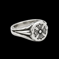 A silver-toned signet-style ring with a textured band and a round face featuring an engraved black floral or cross-like motif, shown on a clean white background.